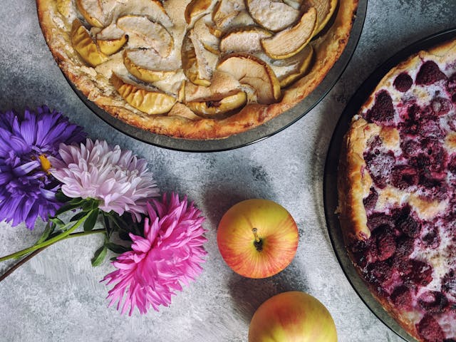 Homemade apple pie with a decorative leaf crust, surrounded by fresh apples and a basket of citrus fruits on a dark wooden table
