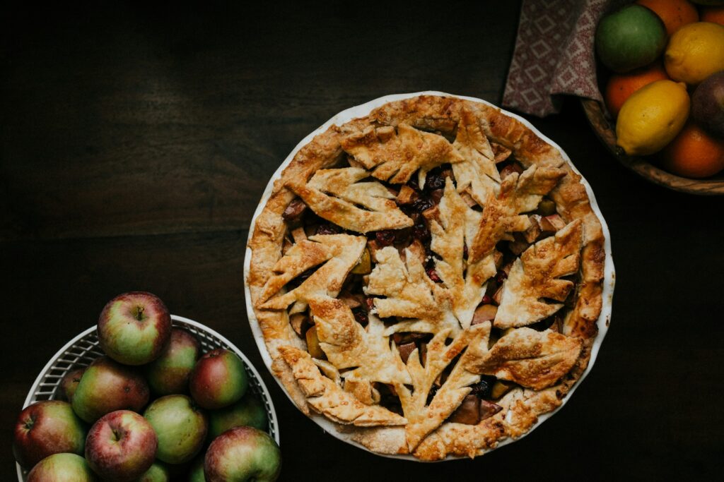 Homemade apple pie with a decorative leaf crust, surrounded by fresh apples and a basket of citrus fruits on a dark wooden table