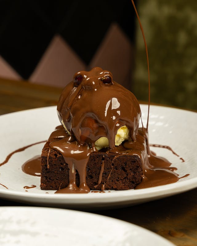 A piece of brownie with pourring chocolate over a white plate.