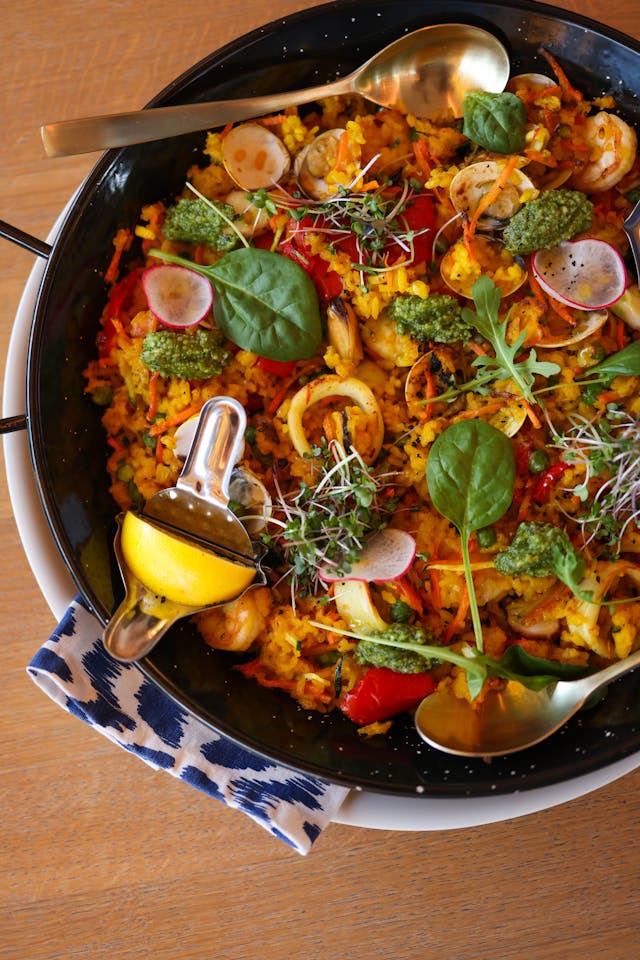 A close-up look on a pan of golden spanish paella with rice, seafood, hearbs, and a slice of lemon.
